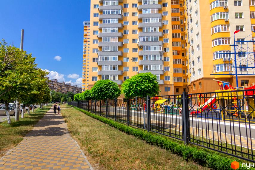 Житловий комплекс на Кондратюка - жовтий багатоповерховий будинок з сірими балконами. Поруч доглянута прибудинкова територія з дитячим майданчиком та зеленими насадженнями.