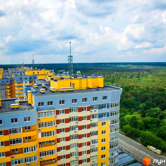 Житловий комплекс на Кондратюка - це багатоповерхові будинки з жовто-сірими фасадами та великою кількістю вікон. Навколо комплексу зелений ліс та дорога з автомобілем.