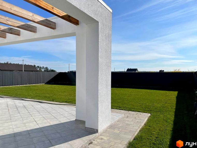 Котеджне містечко КМ Лісне: білий будинок з дерев'яною перголою та доглянутим газоном. Прибудинкова територія з плиткою та зеленим двором.