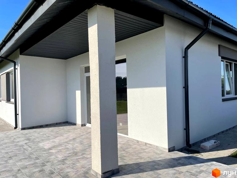 Котеджне містечко КМ Лісне: білий будинок з чорною покрівлею та терасою, вимощеною сірою плиткою. Навколо будинку доглянута прибудинкова територія.