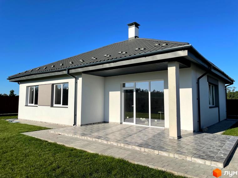 Котеджне містечко КМ Лісне: одноповерховий будинок з сірим дахом, великими вікнами та терасою, оточений зеленою галявиною та вимощеною плиткою прибудинковою територією.