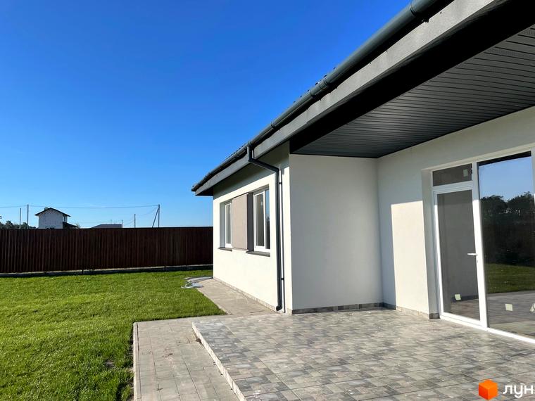 Зовнішній вигляд будинку котеджного містечка КМ Лісне з білою стіною та великими вікнами. Прибудинкова територія з зеленим газоном та вимощеною плиткою терасою.