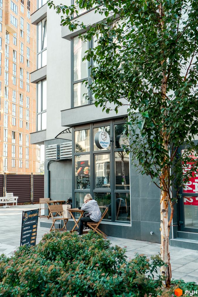 Фасад Житлового комплексу Crystal Avenue з великими вікнами та кафе на першому поверсі з літньою терасою і зеленою зоною.