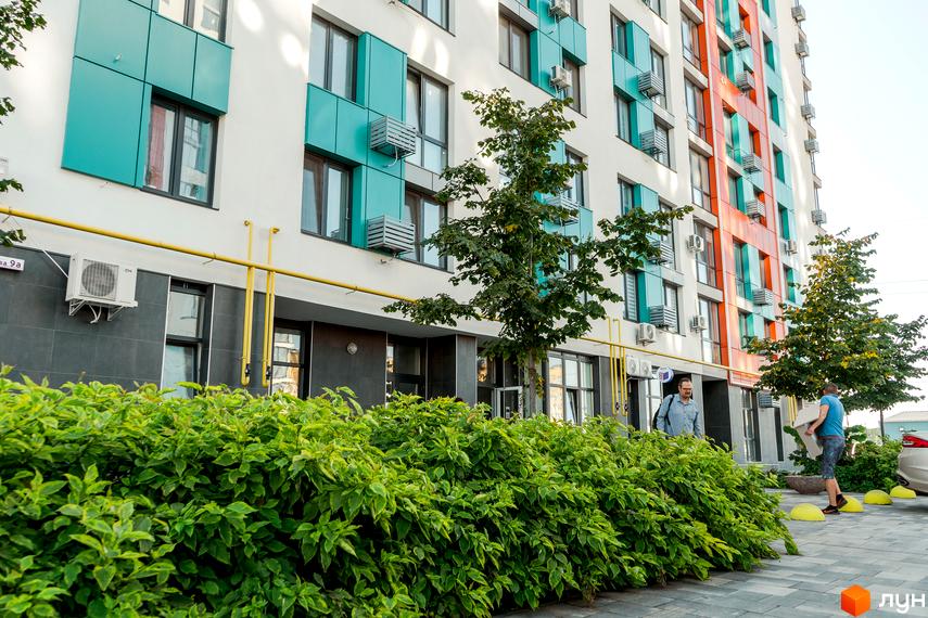 Багатоповерховий будинок житлового комплексу Crystal Avenue з білими стінами, бірюзовими та помаранчевими вставками. Перед будинком зелені кущі та дерева, викладена тротуарна плитка.