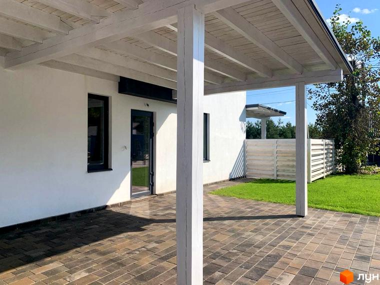 Зовнішній вигляд будинку в КМ Wood Side Village з білою стіною та терасою. Прибудинкова територія з газоном та бруківкою.