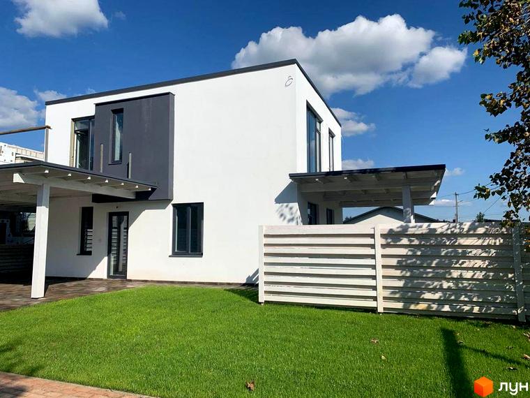 Котеджне містечко Wood Side Village: двоповерховий будинок з білою та сірою фасадною обробкою, навісом та доглянутим газоном. Навколо будинку – зелена галявина та дерев'яний паркан.