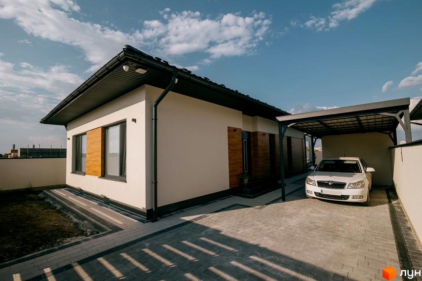 Білий одноповерховий будинок з темним дахом та деревʼяними вставками на фасаді. На території розташована крита парковка та бруківка.