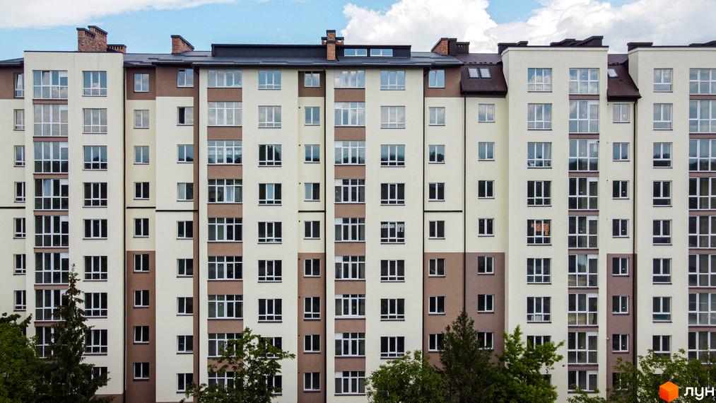 Фасад новобудови ЖК Парковий маєток з численними вікнами та балконами. Будинок має кремово-коричневу кольорову гаму. Навколо видно зелені дерева.