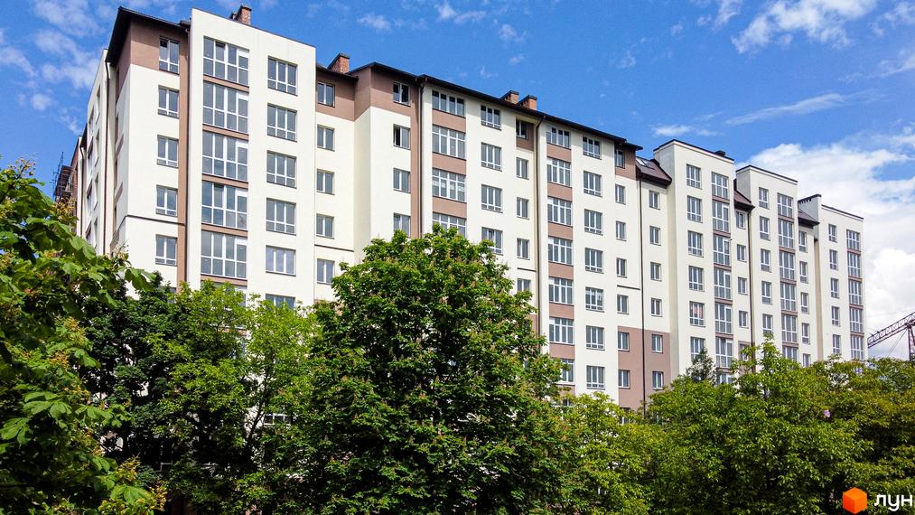 Зовнішній вигляд новобудови ЖК Парковий маєток з численними вікнами та коричневими вставками. Зелені дерева перед будинком.