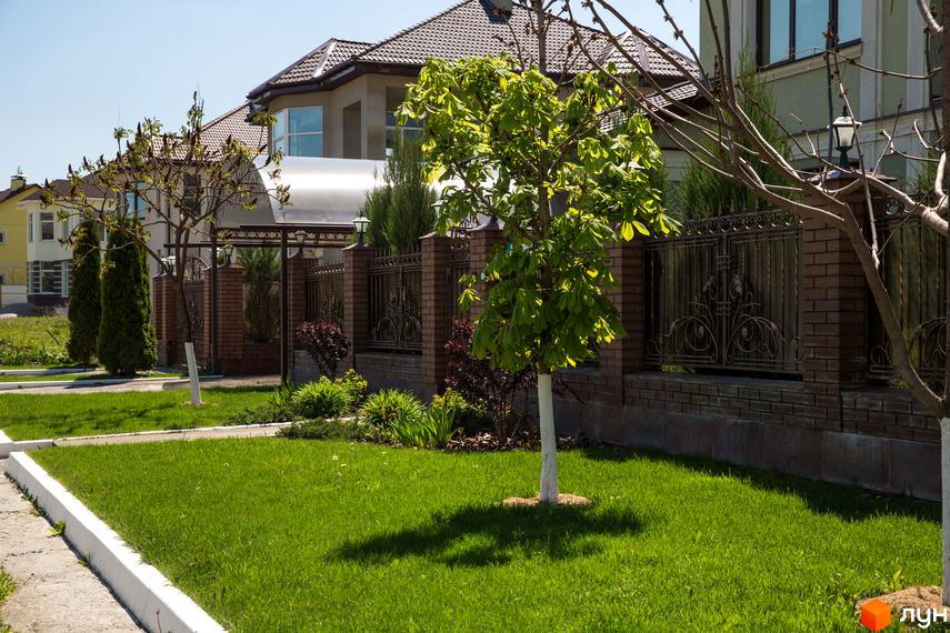 Котеджне містечко КМ Home Resort: фасад будинку з цегляною огорожею та доглянутою зеленою територією. Навколо новобудови висаджені дерева та кущі, газон.