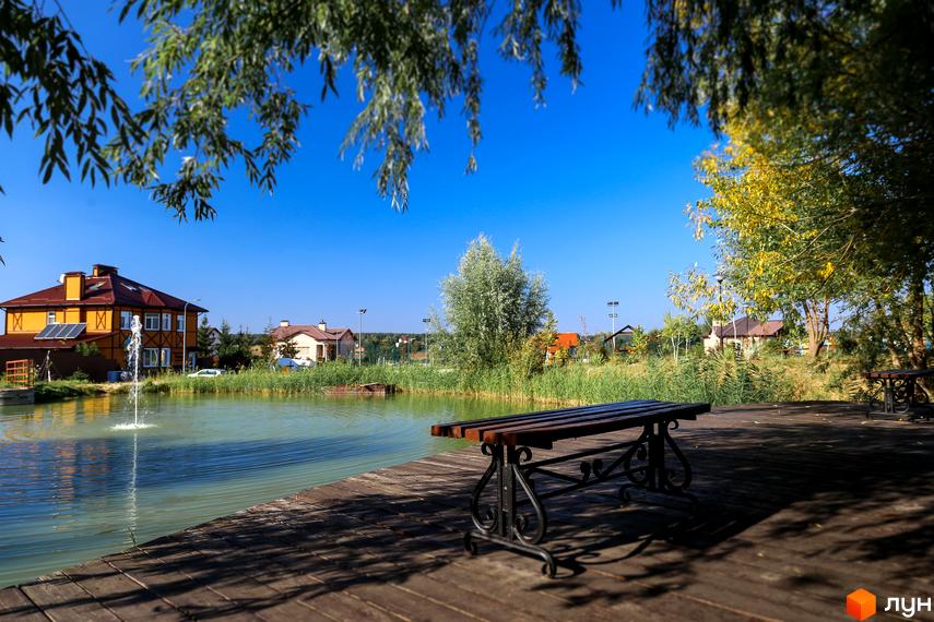 Котеджне містечко КМ Home Resort біля озера з фонтаном. На дерев'яному помості стоїть лавка, навколо зелені дерева та будинки.