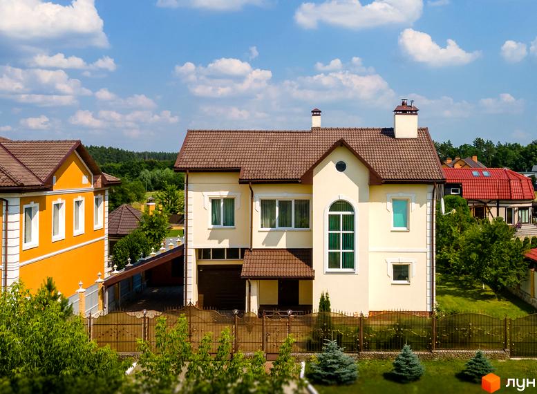 Котеджне містечко КМ Home Resort: двоповерховий будинок світло-бежевого кольору з коричневим дахом та великим арочним вікном. На прибудинковій території зелені насадження та ковані огорожі.