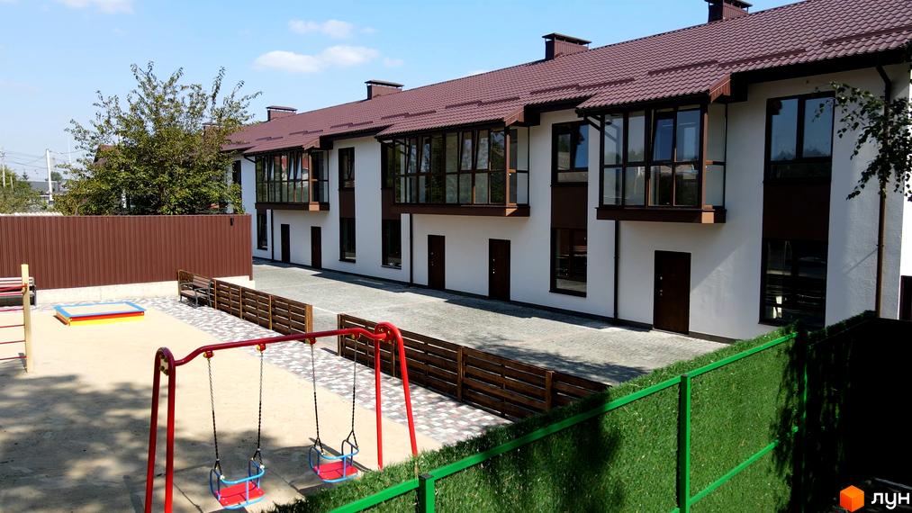 Ряд білих Таунхаусів Green Heaven з коричневими дахами та панорамними вікнами. Перед будинками розташований дитячий майданчик з гойдалками та пісочницею.