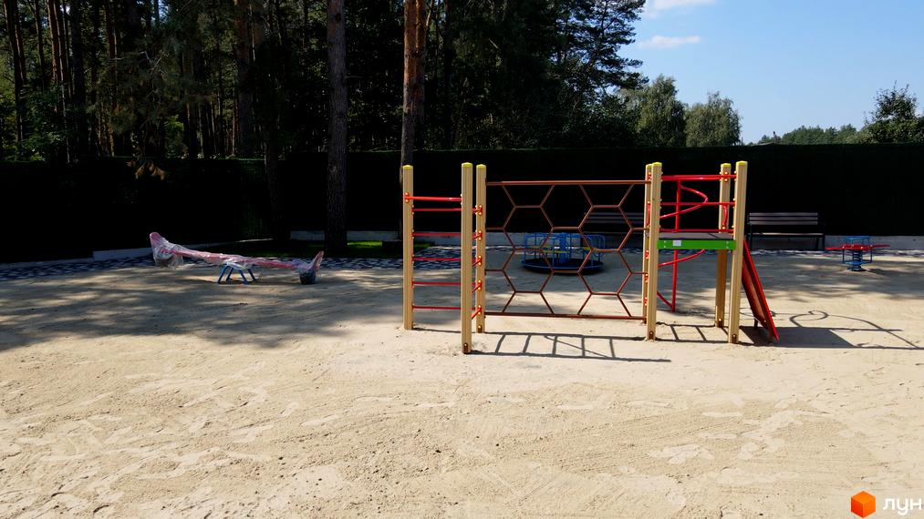 Дитячий майданчик у житловому комплексі Таунхауси Green Heaven з пісочницею, гіркою та гойдалкою. Територія оточена зеленою огорожею та деревами.