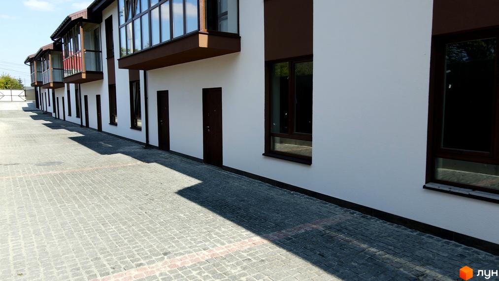 Зовнішній вигляд таунхаусів Green Heaven з білою штукатуркою та коричневими вікнами. Поруч розташований вимощений бруківкою двір.