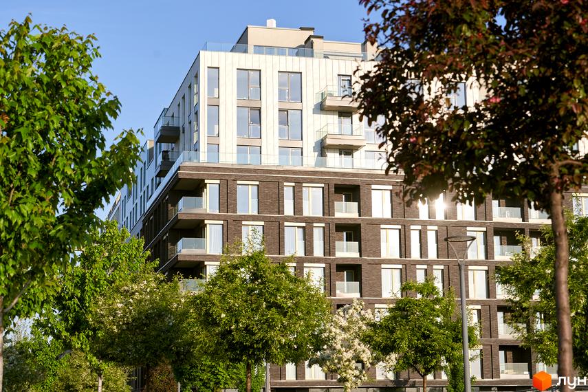 Житловий район Rybalsky: фасади будинку з цегляною обробкою та великими вікнами, балкони зі скляними огорожами. Навколо зелені дерева та блакитне небо.