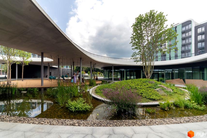 Житловий комплекс Respublika: багатоповерховий будинок з фасадом зеленого кольору та вигнутим бетонним навісом. Під навісом розташований дитячий майданчик, а поруч – водойма з рослинами та острівцем зелені.