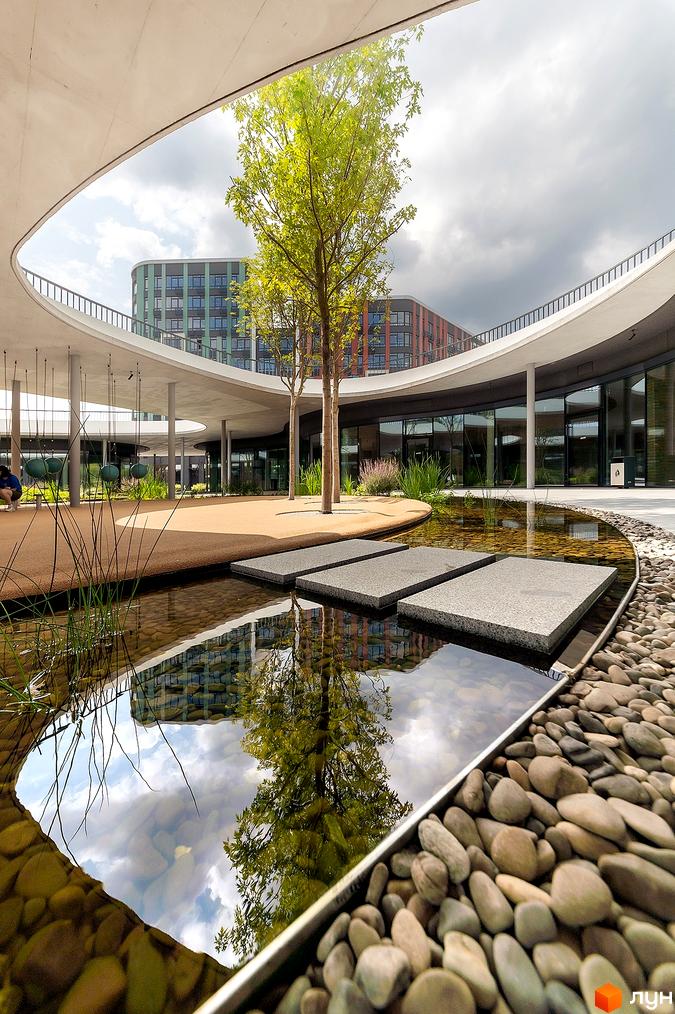 Житловий комплекс Respublika з багатоповерховими будинками з вигнутими фасадами. Прибудинкова територія має водний об'єкт з камінням, зелені дерева та круглий навіс.