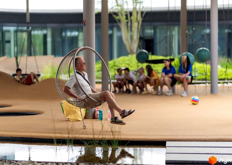 Житловий комплекс Respublika. На прибудинковій території чоловік відпочиває на підвісному кріслі біля водойми, поруч дитячі зони та місця для сидіння.