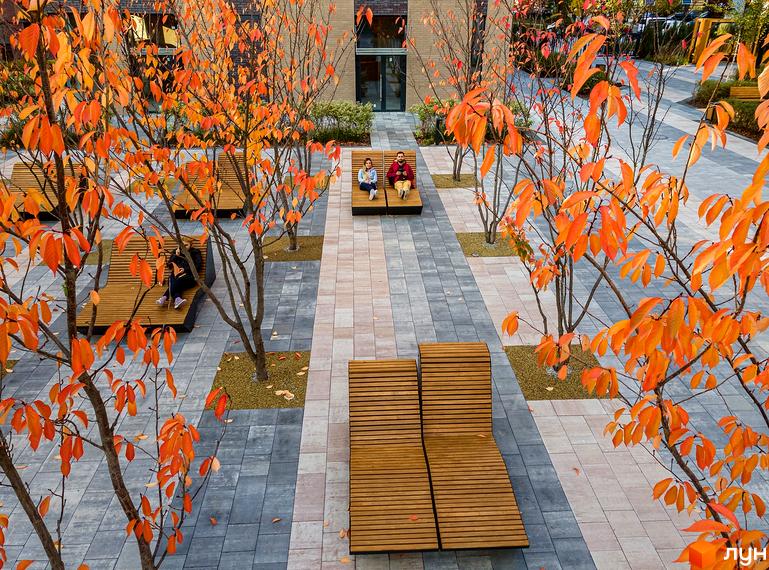 Житловий комплекс Файна Таун з лавками для відпочинку та осінніми деревами з помаранчевим листям. Бруківка та зелені насадження створюють затишну атмосферу.