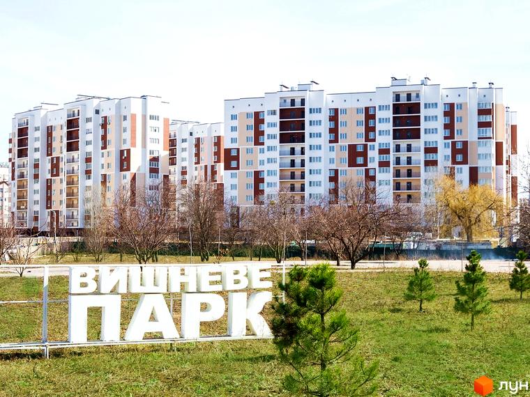 Житловий комплекс Сади Вишневі: багатоповерхові будинки з білими та коричневими фасадами, балконами та великою кількістю вікон. Поруч зелена зона з молодими деревами та написом