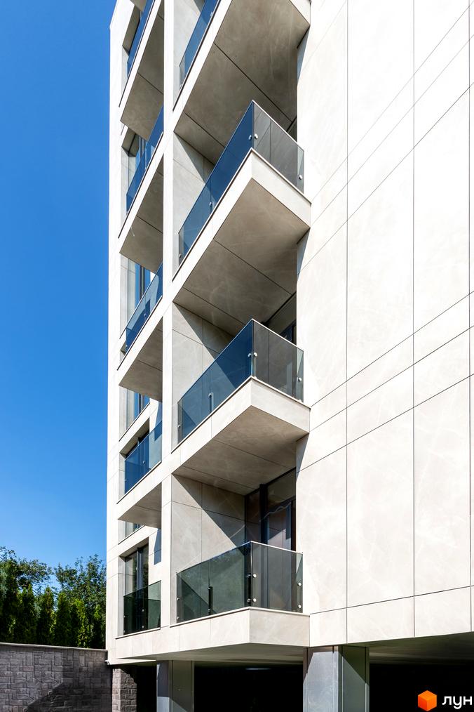 Висотна будівля з балконами зі скляними огорожами. Фасад виконаний у світлих тонах, на тлі блакитного неба.