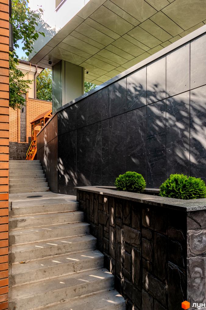 Сходи з бетону ведуть до входу в будівлю. Чорна фасадна плитка та зелені кущі доповнюють архітектуру Клубний будинок Hidden.