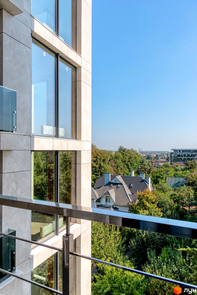 Вид на будівлю житлового комплексу Hidden з балкону. Фасад будинку виконаний у світлих тонах, поруч зелені насадження та приватні будинки.