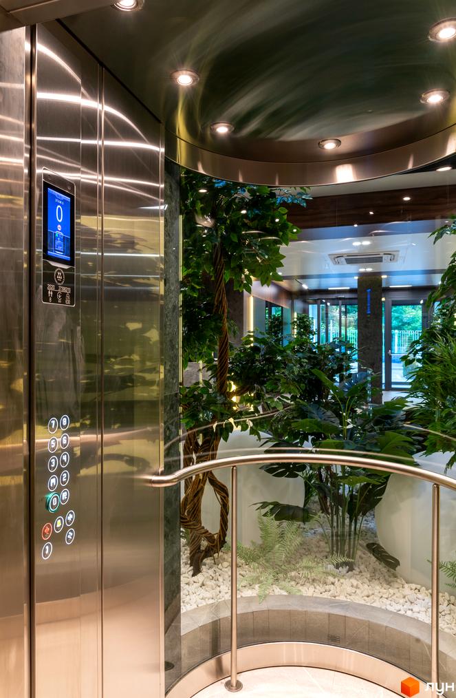 Інтер'єр ліфту в житловому комплексі Hidden з нержавіючої сталі з рослинами та скляними стінами, що відкривають вид на прибудинкову територію.
