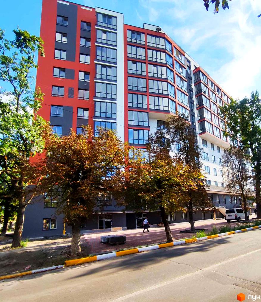 Житловий комплекс Green Life-3 має багатоповерхову будівлю з червоно-сірим фасадом та великою кількістю вікон. Навколо ростуть дерева з осіннім листям.