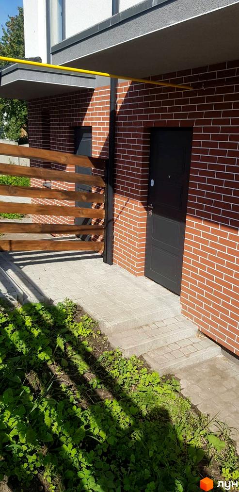 Зовнішній вигляд будинку з червоної цегли з темними дверима та дерев'яною огорожею. Поруч розташована вимощена доріжка та зелені насадження.