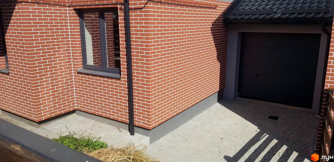 Зовнішній вигляд будинку з червоної цегли з білою розшивкою та гаражними воротами. Прибудинкова територія вимощена плиткою.