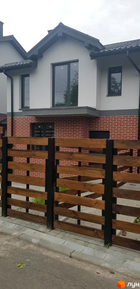 Котеджне містечко КМ Скандинавія: двоповерховий будинок з сірим фасадом, цегляною основою та темними вікнами. Огороджений дерев'яним парканом.