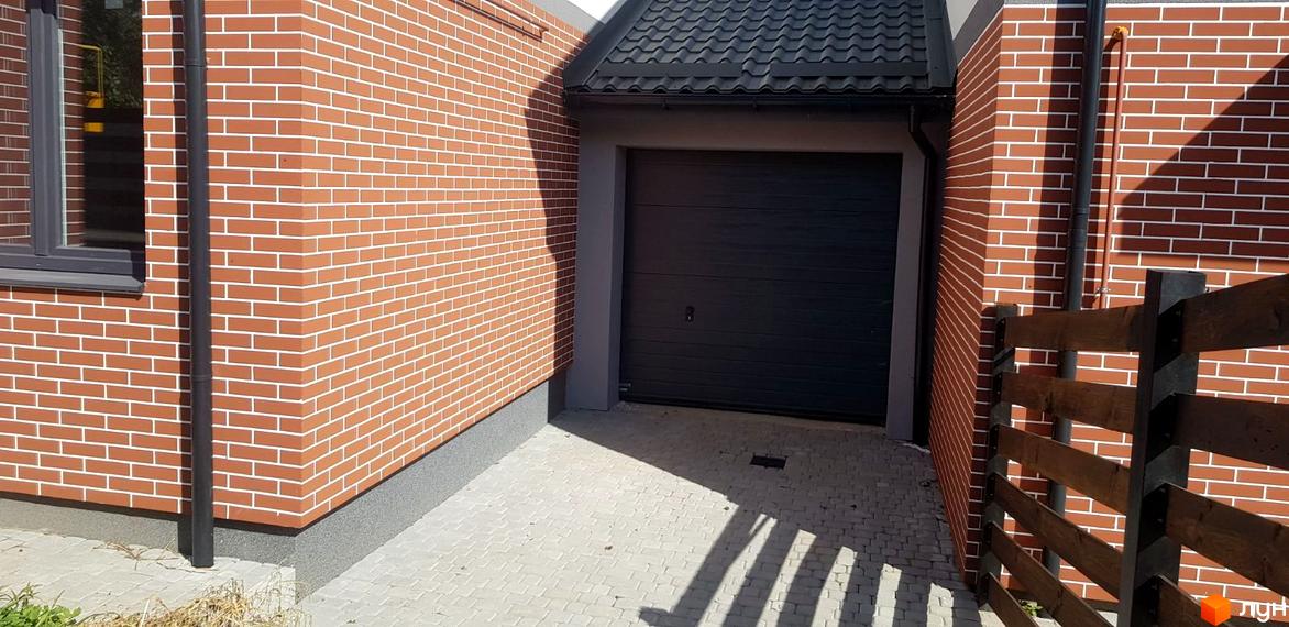 Котеджне містечко КМ Скандинавія: фасад будинку з червоної цегли з гаражними воротами та вимощеною прибудинковою територією. Поруч дерев'яний паркан.