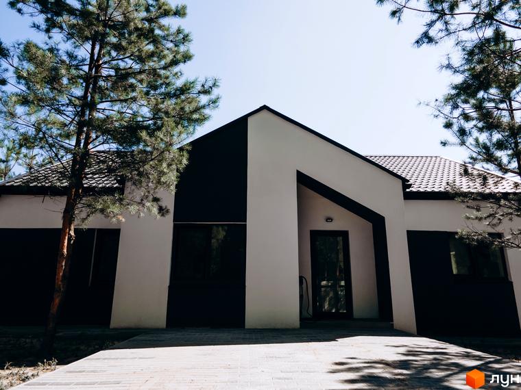 Котеджне містечко Chaletville: фасад будинку з білими та чорними елементами, вхідна група зі скляними дверима, вимощення з плитки. Навколо будинку дерева.