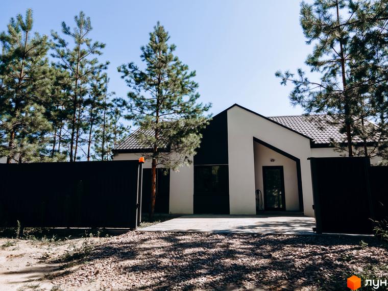 Біло-чорний будинок з темним дахом та вхідною групою на території Котеджного містечка Chaletville. Перед будинком розташована чорна огорожа, вимощена доріжка та високі сосни.