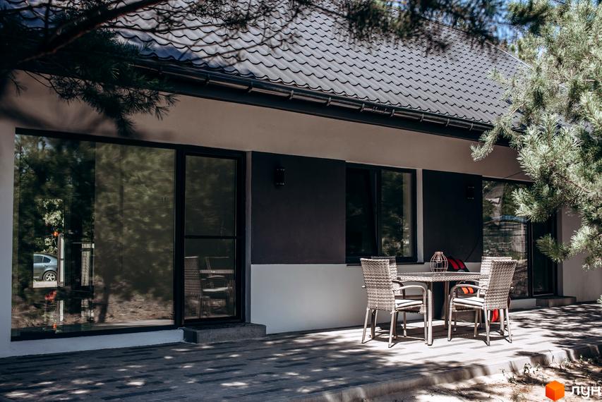 Зовнішній вигляд будинку котеджного містечка КМ Chaletville з терасою та вуличними меблями. Навколо будинку зелені насадження.