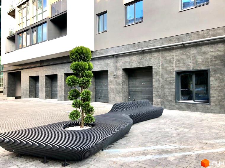 Зовнішній вигляд новобудови ЖК Park Residence з сірою плиткою та великими вікнами. Прибудинкова територія з оригінальною лавкою та деревом.