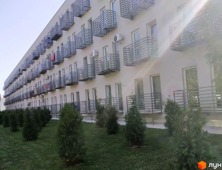 Новобудова Золоті Стовпи з фасадом світлого кольору та численними балконами з металевими огорожами. Перед будинком зелений газон з рядами молодих хвойних дерев.