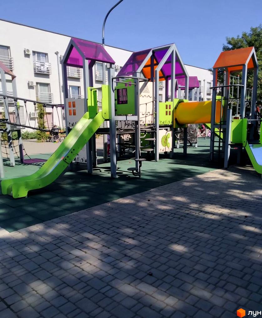 Білий багатоповерховий будинок Житловий комплекс Золоті Стовпи з балконами. На передньому плані дитячий майданчик з зеленими гірками та жовтим тунелем.