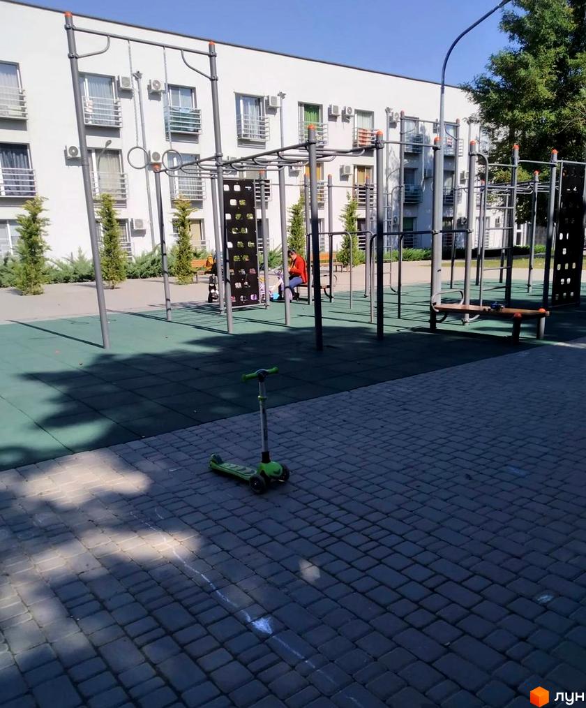 Білий багатоповерховий будинок Комплексу Золоті Стовпи з численними балконами. Поруч облаштований спортивний майданчик з тренажерами та зеленим покриттям.