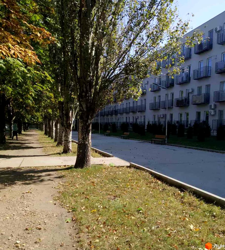 Житловий комплекс Золоті Стовпи: багатоповерховий будинок зі світлим фасадом та балконами. Прилегла територія з алеєю дерев, пішохідною доріжкою та лавками.