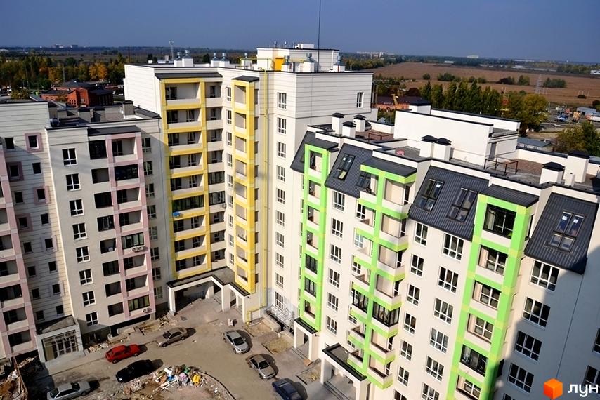 Житловий комплекс Акварелі: багатоповерхові будинки світлих відтінків з жовтими, рожевими та зеленими елементами фасаду. Прибудинкова територія з автомобілями та будівельними матеріалами.