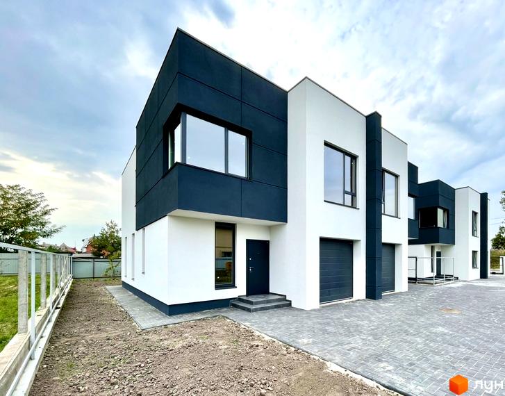 Ряд двоповерхових таунхаусів з білим та темно-сірим фасадом, великими вікнами та гаражами. Перед будинками бруківка, поруч ділянка з ґрунтом.