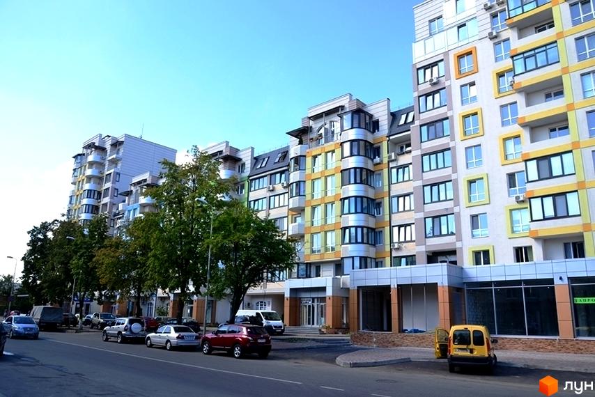 Житловий комплекс Акварелі: багатоповерхові будинки з різнокольоровими фасадами та округлими балконами, вздовж вулиці припарковані автомобілі та ростуть дерева.