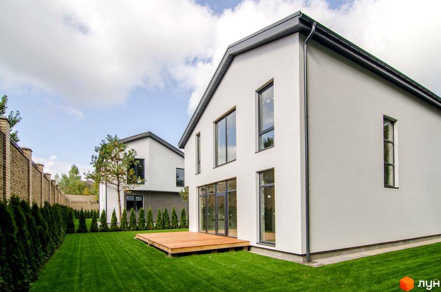 Котеджне містечко КМ Friends Club. Білий двоповерховий будинок з великими вікнами та дерев'яною терасою, зелений газон та огорожа з туй.