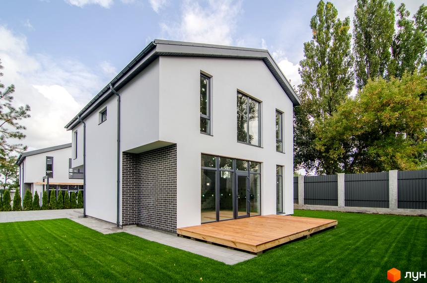 Котеджне містечко КМ Friends Club: двоповерховий будинок з білою штукатуркою, сірою цеглою та дерев'яною терасою. Доглянутий зелений газон та паркан.