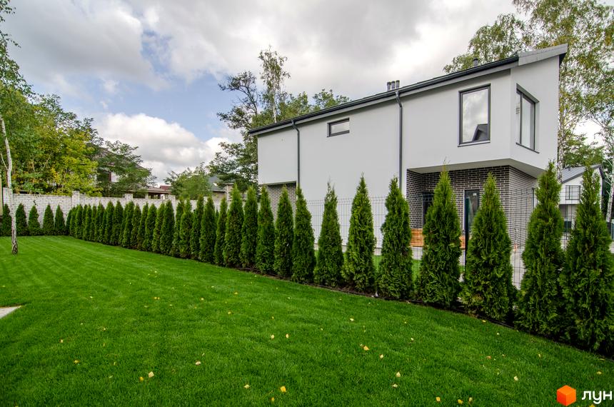 Котеджне містечко КМ Friends Club: двоповерховий будинок з білою штукатуркою, цегляною нижньою частиною та зеленою галявиною з туями.