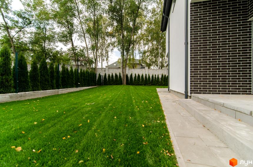 Котеджне містечко Friends Club: фасад будинку з темної цегли та білої штукатурки, зелений доглянутий газон та туї вздовж паркану.