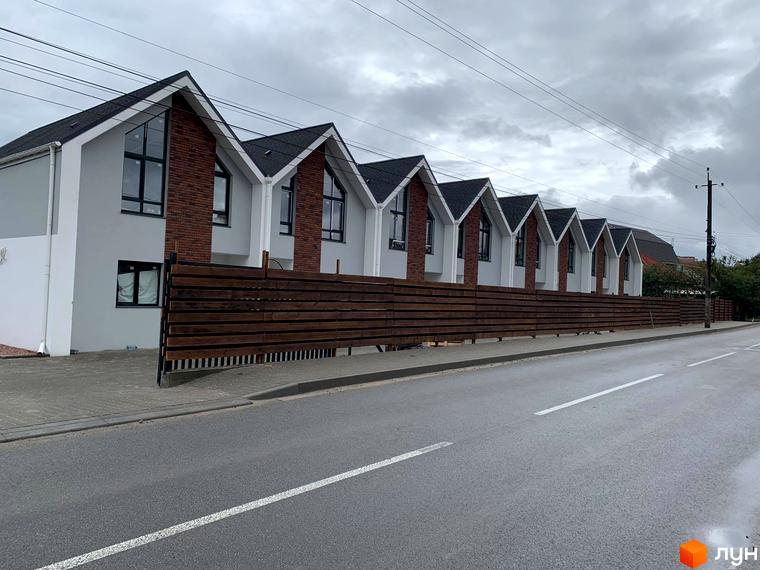 Зовнішній вигляд таунхаусів Scandi House з цегляними вставками та темним дахом, розташованих уздовж дороги з дерев'яною огорожею.
